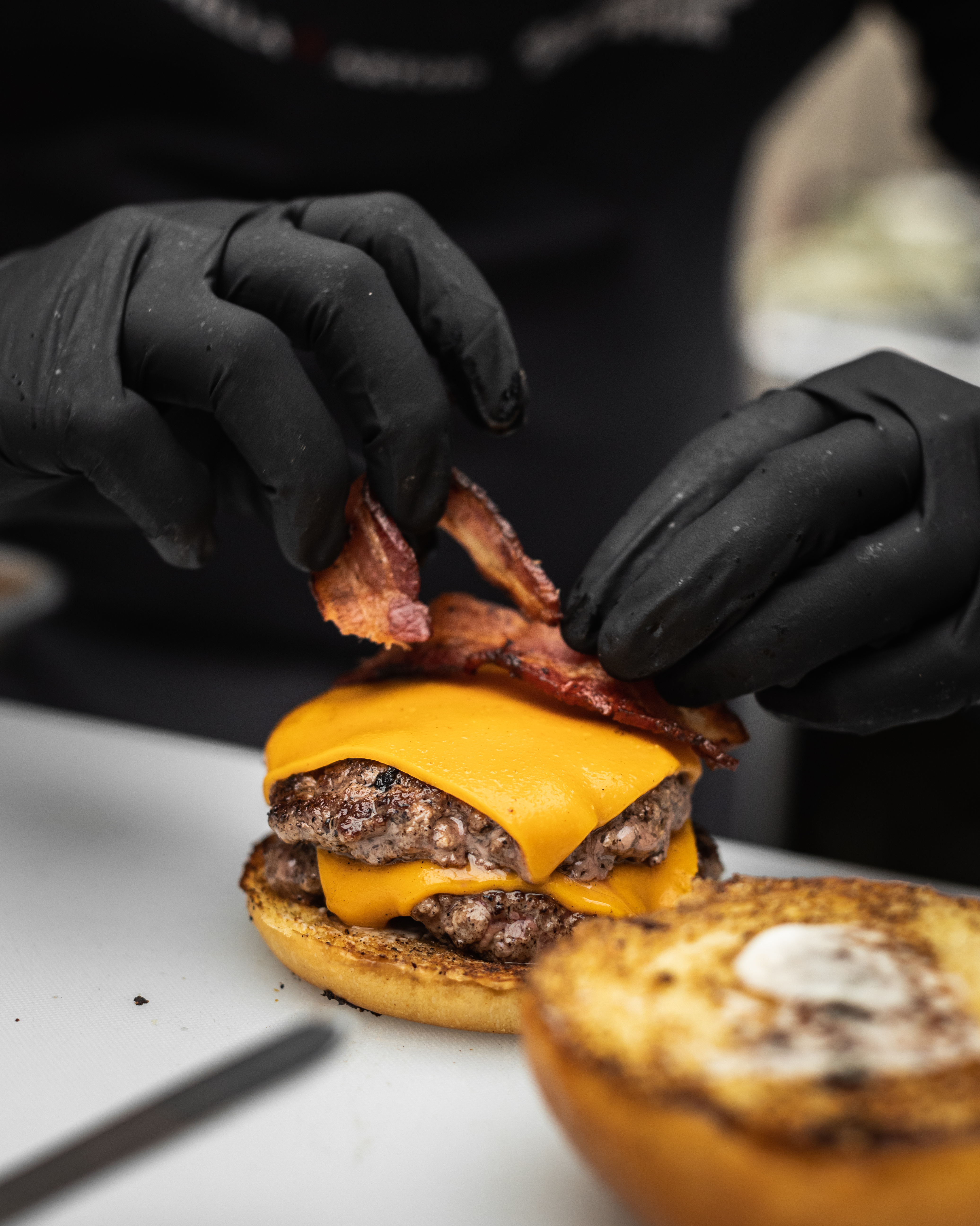 Imagem de duas mãos com luvas pretas, preparando um Hamburguer